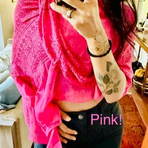 Può includere: Uno scialle rosa acceso caratterizzato da un motivo scuro, intricato e ripetitivo. Il tessuto è drappeggiato sul busto e presenta una consistenza leggera. Il testo "Pink!" e "Use as a wrap, shawl, cover, or scarf" è stampato in rosa in basso.