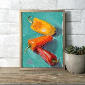 Pode incluir: Uma pintura emoldurada de três pimentões em um fundo turquesa. Os pimentões são vermelho, laranja e amarelo. A pintura está em uma moldura dourada.