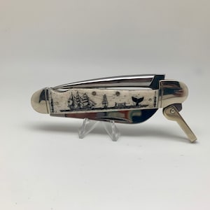May include: A folding pocket knife with a cream-colored handle featuring a black etched illustration of a sailing ship, a smaller boat with figures, and a whale tail. The knife has polished silver-tone metal bolsters and a metallic blade edge.
