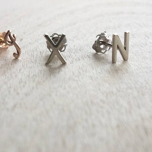 May include: Three pairs of earrings with initial letters L, X, and N. The L earring is gold, the X earring is silver, and the N earring is silver.
