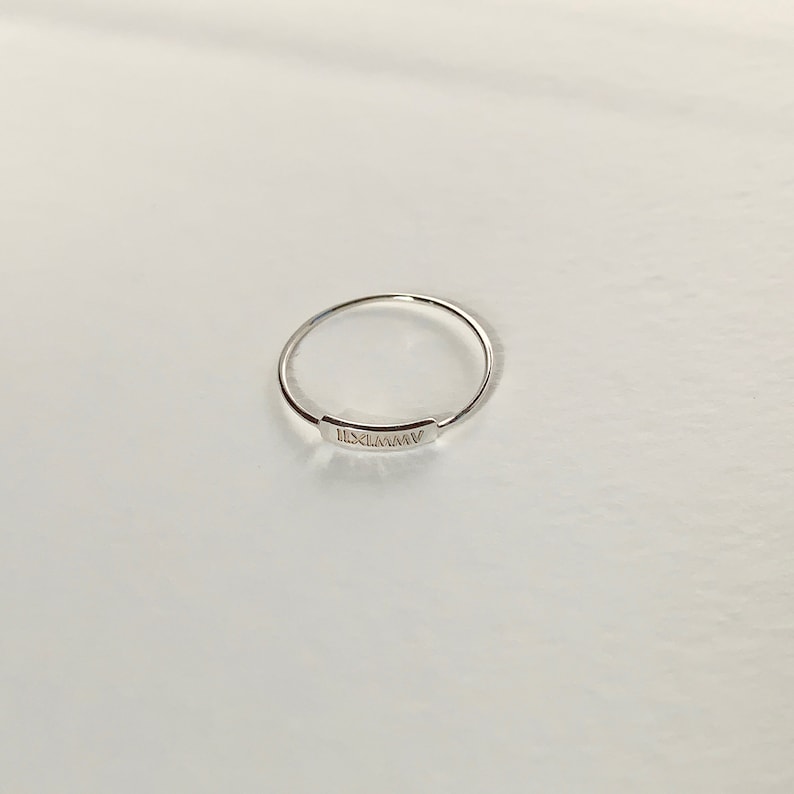 May include: A silver ring with a thin band and a rectangular plate engraved with the Roman numeral 'IXLMMIV'.