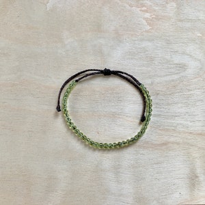 May include: A green gemstone beaded bracelet with a brown cord. The bracelet is tied with a knot and is ready to be worn.