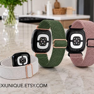 May include: Three Fitbit smartwatches with elastic bands in light gray, olive green, and dusty pink. Each watch features a black casing, a sensor array on the back, and a metal slider for size adjustment. The text ALEXIUNIQUE.ETSY.COM is visible.