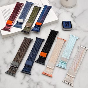 May include: A collection of ten woven nylon watch straps in various colours including maroon, blue, olive green, black, beige, light blue, and white. Each strap features a metal connector and a small rectangular pull tab in contrasting colours.
