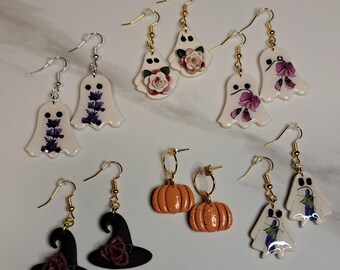 May include: A collection of handmade Halloween-themed earrings. The earrings feature ghost shapes with floral designs, witch hats with roses, and orange pumpkin shapes. The earrings have gold and silver hooks.