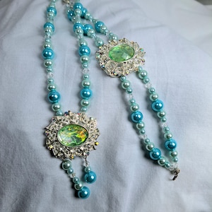 May include: Two beaded necklaces with a vintage aesthetic. Each necklace features a large, ornate silver-toned pendant with a green oval-shaped stone. The necklaces are strung with light blue and clear beads, with dangling blue bead accents.