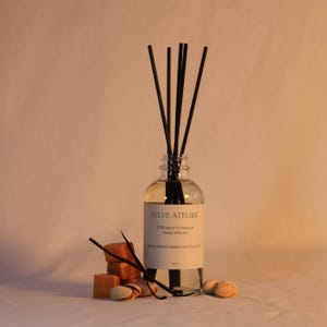 Peut inclure: Un diffuseur de roseaux en verre transparent avec des roseaux noirs, étiqueté "SYLVE ATELIER". La bouteille contient un liquide de couleur claire. Des petits savons carrés brun-orangé et quelques pistaches accompagnent le diffuseur.