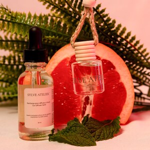 May include: A clear glass bottle with a dropper and a small car air freshener hanging by a string. The bottle has a label that reads "SYLVE ATELIER". A grapefruit slice and mint leaves are in the background.