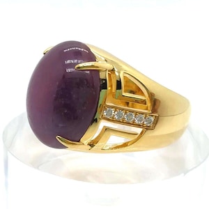 May include: A gold ring featuring a large, oval, deep purple gemstone. The ring has a decorative gold setting with small, clear stones and a polished finish. The ring is displayed on a clear stand.