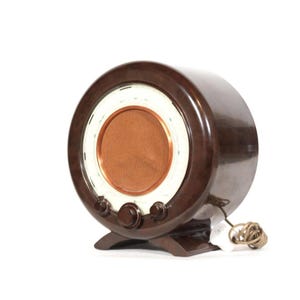 Puede incluir: Una radio circular vintage marrón con una cubierta de altavoz de color cobre y detalles en blanco. La radio tiene tres perillas y un cable de alimentación. El diseño recuerda la estética moderna de mediados de siglo.