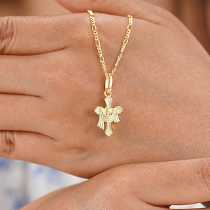 May include: A gold-colored pendant necklace featuring a cross design. The cross has a detailed, ornate appearance with draped elements. The necklace chain is a delicate figaro style, and the pendant is held by a small bail.