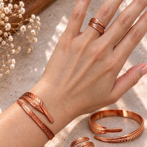 Peut inclure: Un ensemble de bijoux couleur cuivre avec un motif inspiré d'une plume. La collection comprend un bracelet manchette de style enveloppant porté au poignet, une bague ouverte assortie au doigt, ainsi qu'un bracelet et une bague supplémentaires.