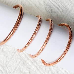 May include: Four copper-coloured metal bangles arranged in a row. The first bangle is smooth and plain. The second features a star-like etched pattern. The third has a textured, cross-hatched surface. The fourth bangle displays a diagonal twisted design.