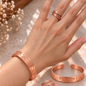 May include: A copper jewelry set consisting of two cuff bracelets and two rings. The bracelets feature an embossed floral vine pattern. One ring has a textured feather-like design, while the other ring is a simple band. The metal has a warm, metallic finish.