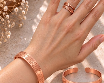 Pode incluir: Um conjunto de joias de cobre composto por dois braceletes e dois anéis. Os braceletes apresentam um padrão floral em relevo. Um anel tem um design texturizado semelhante a uma pena, enquanto o outro anel é uma banda simples de metal.