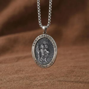 May include: Silver oval pendant necklace depicting Saint Christopher. The pendant shows Saint Christopher carrying a child, with the words "SAINT CHRISTOPHER PROTECT US" around the edge. The necklace has a chain.