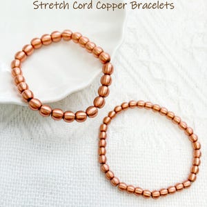Könnte beinhalten: Zwei Armbänder aus Kupfer auf elastischem Band mit glatten, runden, metallisch kupferfarbenen Perlen. Der Text "Stretch Cord Copper Bracelets" ist oben im Bild zu sehen. Die Armbänder sind kreisförmig auf einer weißen Oberfläche angeordnet.