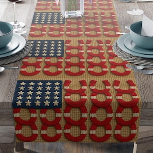 Puede incluir: Un camino de mesa patriótico con un diseño que recuerda a la bandera estadounidense. El camino presenta cuadrados azul marino con estrellas doradas y patrones de anillos rojos y blancos sobre un fondo beige. La mesa está puesta con vajilla, cubertería y copas de vino.