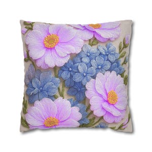 Puede incluir: Un cojín cuadrado con un intrincado diseño bordado sobre tela de color beige claro. El patrón muestra grandes flores de color violeta claro con centros amarillos, racimos de pequeñas flores azules y tallos verdes con hojas.
