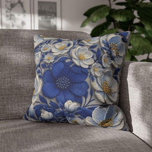 May include: Square throw pillow featuring a floral pattern with large, deep blue flowers and smaller white and light blue blossoms. The pillow has a gray background and is placed on a gray sofa.