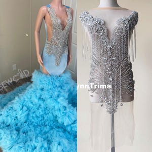 May include: A beaded applique with a sheer fabric backing. The applique features a V-neckline and a cascading design of rhinestones and beads. The applique is shown on a mannequin.