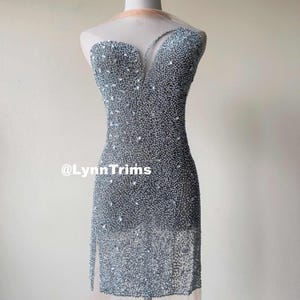 May include: A silver sequined dress with a sheer overlay. The dress has a V-neckline and a fitted silhouette. The dress is embellished with rhinestones and beads. The dress is shown on a mannequin. The text "@LynnTrims" is visible on the dress.