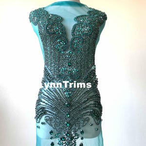 Könnte beinhalten: Eine tealgrüne Perlenverzierung mit einem V-Ausschnitt. Die Verzierung befindet sich auf einem durchsichtigen Stoffhintergrund. Der Text "LynnTrims" ist unten in der Mitte des Bildes sichtbar.