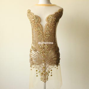 May include: A gold-colored beaded applique with an intricate design. The applique features a central pattern with a fan-like lower section and a decorative neckline. The design is embellished with various sizes of gold beads and crystals. The text "@LynnTrims" is visible.