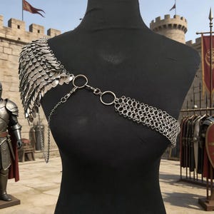 May include: Silver shoulder armour with a scale design and chainmail across the chest, displayed on a black mannequin. A chain connects the shoulder piece to the chest piece. The background features a castle and other armour.