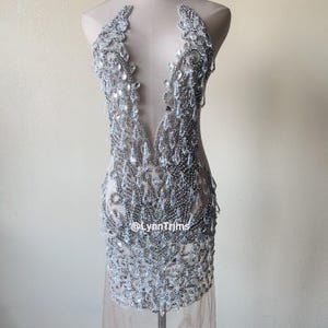 May include: A silver beaded and sequined dress with a plunging neckline and a sheer overlay. The dress is on a mannequin and has a beaded design that resembles teardrops. The text "@LynnTrims" is visible on the dress.