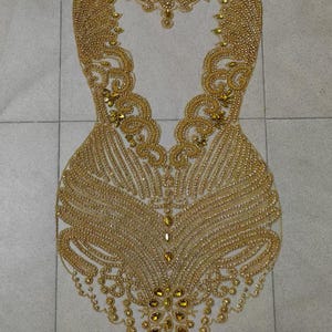 May include: A gold beaded embellishment with a delicate, intricate design. The embellishment is shaped like a bodice and features a variety of bead sizes and shapes.