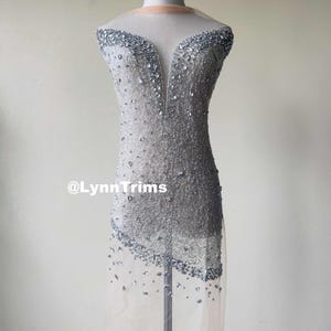 May include: A silver beaded fabric with a sheer overlay. The fabric is embellished with rhinestones and has a V-neckline. The fabric is shown on a dress form. The text "@LynnTrims" is visible in the lower left corner of the image.