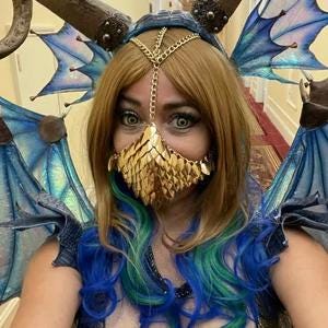 May include: A woman wearing a gold chain headpiece and a gold scale mask with blue and green hair. She is wearing a blue and iridescent costume with large wings.
