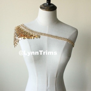 May include: Gold chain shoulder piece with a cascading design of gold metal leaves. The chain is worn on a white mannequin. The text '@LynnTrims' is visible in the image.