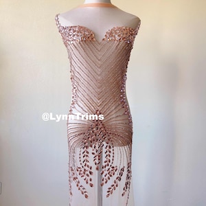 Może przedstawiać: Różowo-złota aplikacja z koralików do ubrań. Aplikacja wykonana jest z małych, fasetowanych koralików, które są przyszyte do przezroczystej siateczki. Aplikacja jest przeznaczona do przyszycia do odzieży, takiej jak sukienka lub bluzka, aby dodać jej blasku i elegancji.