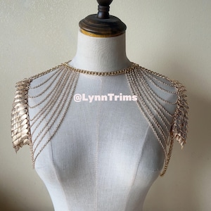 以下が含まれることがあります： チェーンと鱗のディテールが施されたゴールドトーンのショルダーアクセサリー。マネキンに飾られており、肩から流れ落ちる繊細なチェーンが複数あります。テキスト「@LynnTrims」が表示されています。