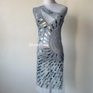 May include: A silver sequined and mirrored fabric applique for a dress or other garment. The applique features a geometric design with a circular motif. The fabric is sheer and has a delicate texture.
