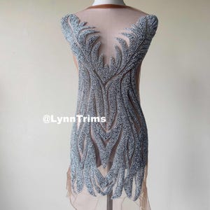 Puede incluir: Un diseño de aplique de cuentas plateadas sobre una tela nude transparente. El diseño presenta un patrón simétrico de hojas y ramas.