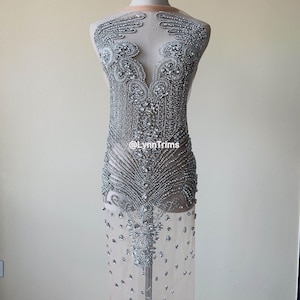Könnte beinhalten: Eine silberne Perlenverzierung für ein Kleid oder ein Kleidungsstück. Die Verzierung besteht aus kleinen, runden Perlen, die in einem komplizierten Muster zusammengenäht sind. Die Verzierung ist zum Annähen an ein Kleidungsstück gedacht und kann verwendet werden, um jedem Outfit einen Hauch von Glanz und Glamour zu verleihen.