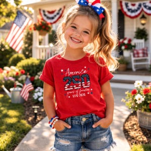 May include: A young person wearing a red t-shirt with the text "America 250 years of freedom 1776-2026" in blue and white. The person is also wearing denim shorts, a red, white, and blue bow, and a bracelet. American flags and patriotic decorations are in the background.