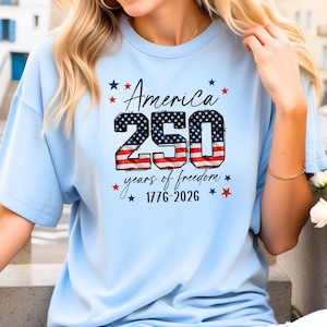 May include: Light blue t-shirt with the text "America 250 years of freedom 1776-2026" in a patriotic design. The number 250 is filled with a stars and stripes pattern, and the text is surrounded by red and blue stars.