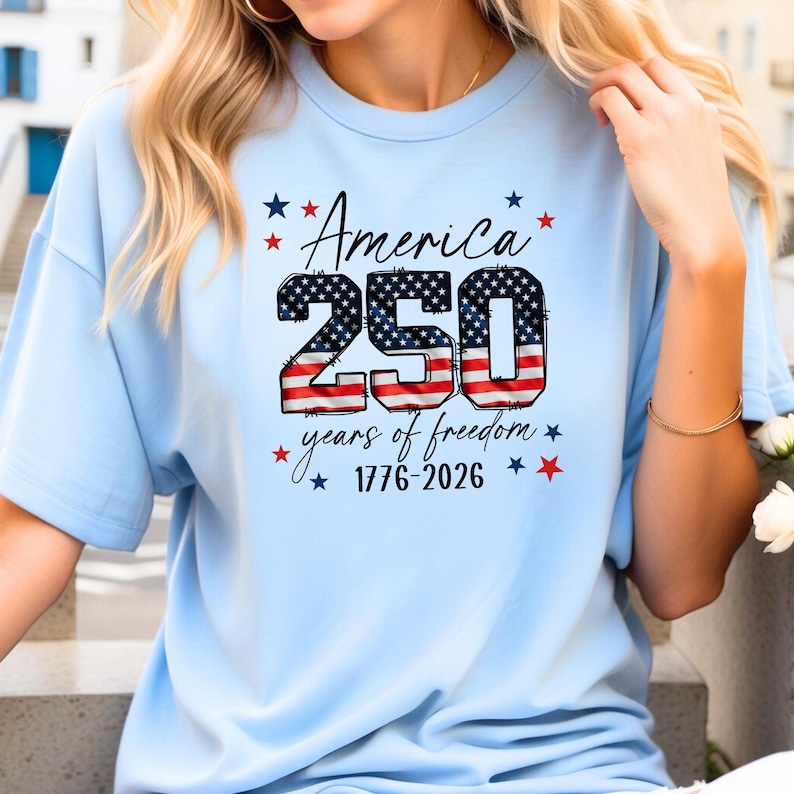 May include: Light blue t-shirt with the text "America 250 years of freedom 1776-2026" in a patriotic design. The number 250 is filled with a stars and stripes pattern, and the text is surrounded by red and blue stars.