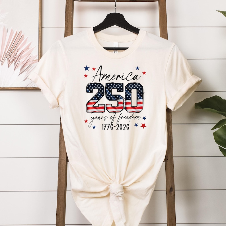 May include: Cream-colored t-shirt featuring the text "America 250 years of freedom 1776-2026" in red, white, and blue. The number "250" is designed with a flag pattern. The shirt is hanging on a black hanger.