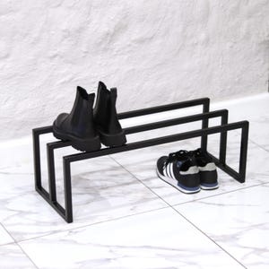 Peut inclure: Un présentoir à chaussures en métal noir avec deux paires de chaussures. La tablette supérieure présente des bottines Chelsea noires. La tablette inférieure présente des baskets noires et blanches avec des détails bleus. Le présentoir est sur un sol carrelé blanc.