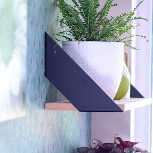 Op de afbeelding: Een zwarte metalen plankbeugel met een houten plank. Een witte keramische pot met een groene plant staat op de plank.