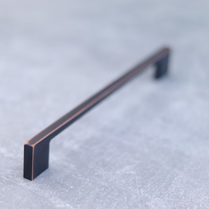 May include: A long, rectangular cabinet handle with a dark matte black finish and subtle copper-toned edges. The hardware features a straight, linear design with two perpendicular mounting posts at each end, creating a clean, geometric profile.