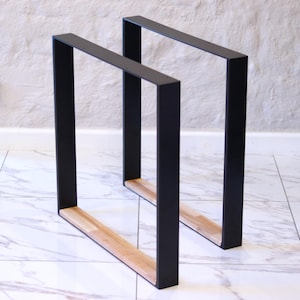 Puede incluir: Dos patas de mesa de metal negro con diseño rectangular. Las patas tienen una inserción de madera natural en la base. Las patas están diseñadas para soportar una mesa. Fondo blanco y suelo de baldosas de mármol.
