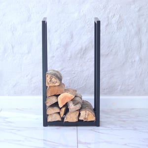 May include: A tall, rectangular black metal log holder with a U-shaped frame. The holder is filled with a stack of split, light brown logs with textured bark, arranged in a tiered formation within the vertical metal supports.