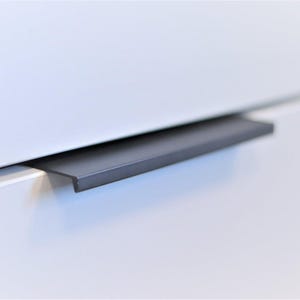 Puede incluir: Primer plano de un tirador de armario negro mate, de forma rectangular y montado horizontalmente en una puerta de armario blanca. El diseño es minimalista y elegante, adecuado para la decoración del hogar contemporánea.