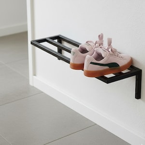 Op de afbeelding: Een zwart metalen schoenenrek aan een witte muur, met een paar roze suède sneakers. De sneakers hebben bruine zolen en groene accenten. Het rek heeft een eenvoudig, geometrisch ontwerp.